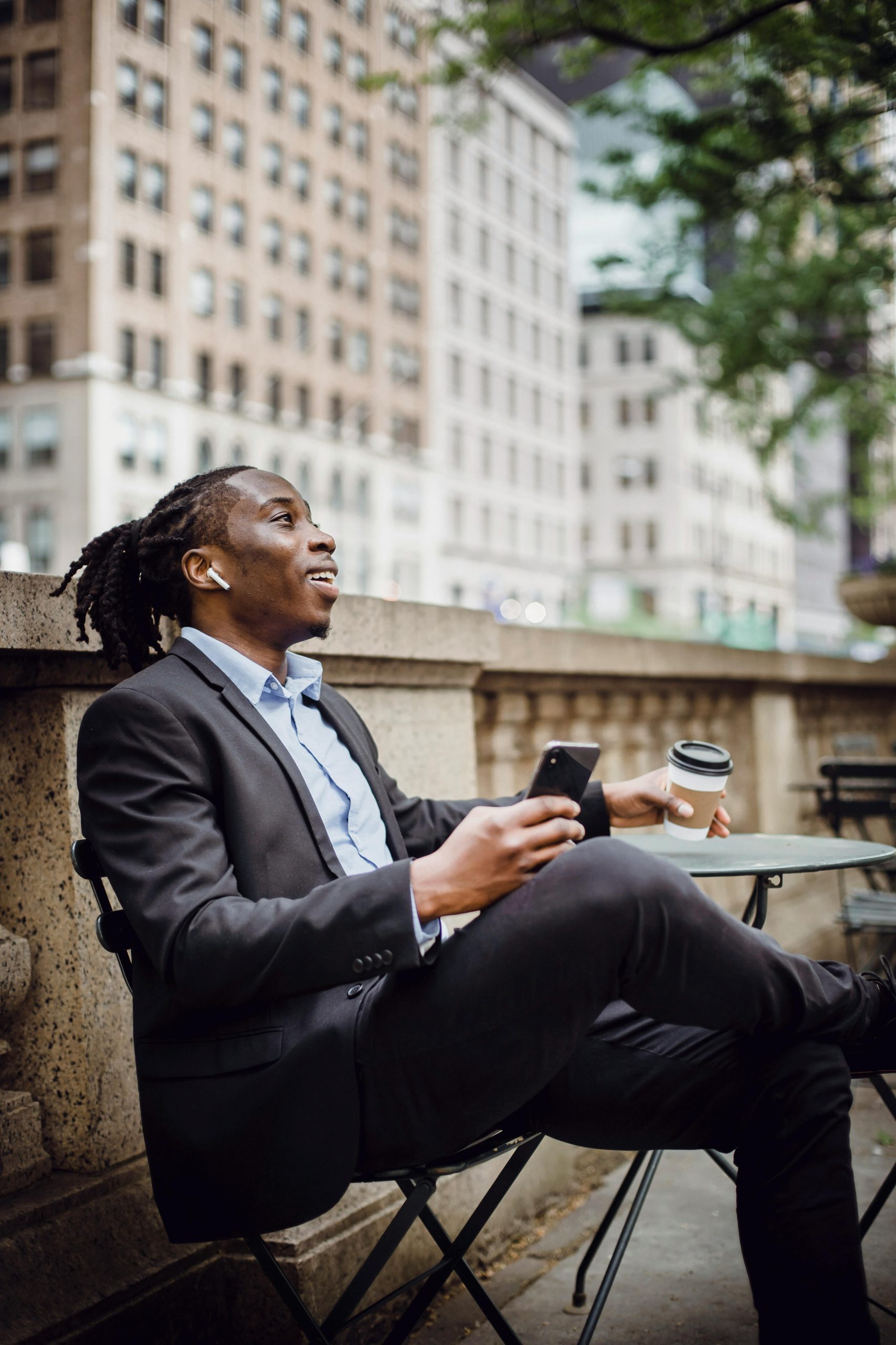 Side view of happy young African American male manager in suit relaxing on terrace of cafe with smartphone and cup of takeaway coffee